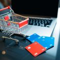 Coș de cumpărături, laptop și carduri financiare pe o masă.